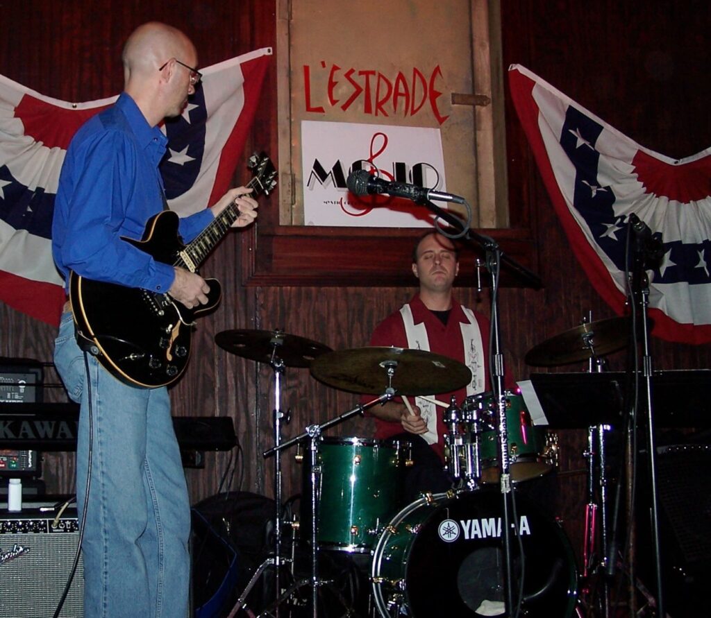 A man playing guitar and drums in front of another man.